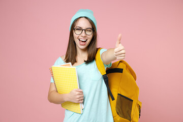 Blinking young woman student in blue t-shirt hat glasses backpack hold notebooks showing thumb up...