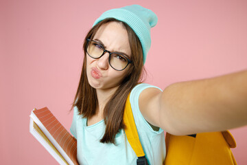 Close up confused woman student in blue t-shirt hat glasses backpack hold notebooks doing selfie shot on mobile phone isolated on pink background. Education in high school university college concept.