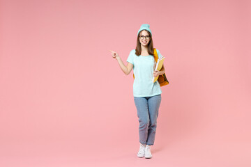 Fototapeta na wymiar Full length of cheerful young woman student in blue t-shirt hat glasses backpack hold notebooks point finger aside isolated on pink background. Education in high school university college concept.