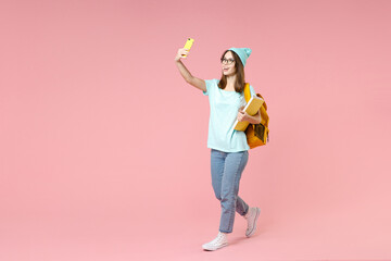 Full length side view of funny woman student in t-shirt hat glasses backpack hold notebooks doing selfie shot on mobile phone isolated on pink background. Education in high school university concept.