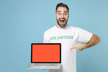 Excited young man in volunteer t-shirt point index finger on laptop pc computer with blank empty screen isolated on blue background studio. Voluntary free work assistance help charity grace concept.