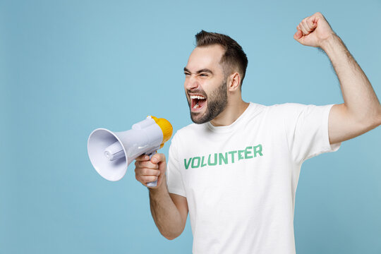Crazy young man wearing white volunteer t-shirt screaming in megaphone clenching fist isolated on blue color background studio portrait. Voluntary free work assistance help charity grace concept.