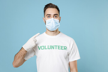 Young man in volunteer t-shirt sterile gloves pointing finger on face mask to safe from coronavirus virus covid-19 isolated on blue background. Voluntary free work assistance aid help support concept.