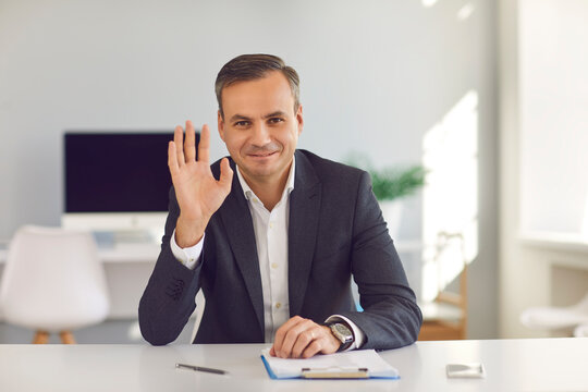 Happy smiling man in suit sitting at office desk, looking at camera and waving hand saying hello during video call. Webcam portrait of business coach, CEO, financial consultant or university professor