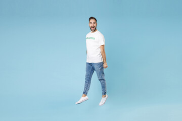 Full length of excited funny young bearded man 20s wearing white volunteer t-shirt jumping isolated on blue color background studio portrait. Voluntary free work assistance help charity grace concept.
