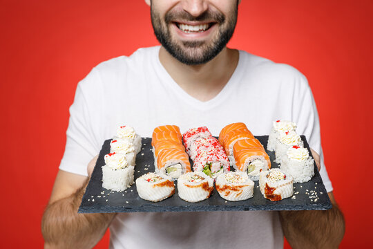 Cropped image of smiling young bearded man 20s in casual white t-shirt hold in hands makizushi sushi roll served on black plate traditional japanese food isolated on red background studio portrait.