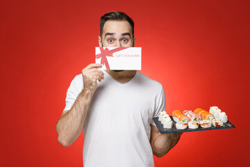 Funny young bearded man 20s in white t-shirt covering face with gift certificate makizushi sushi roll served on black plate traditional japanese food isolated on red color background studio portrait.