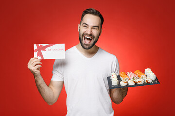 Overjoyed young bearded man 20s in white t-shirt hold in hand gift certificate makizushi sushi roll served on black plate traditional japanese food isolated on red color background studio portrait.