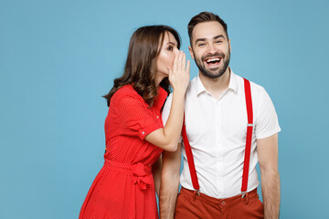 Funny young couple friends man woman in white red clothes whispering gossip tells secret behind her hand, sharing news isolated on blue background studio portrait. St. Valentine's Day holiday concept.