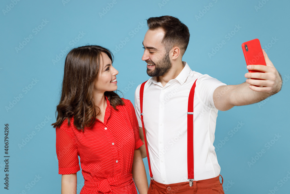 Wall mural Funny young couple two friends man woman in white red clothes doing selfie shot on mobile phone looking at each other isolated on blue background studio portrait. St. Valentine's Day holiday concept.