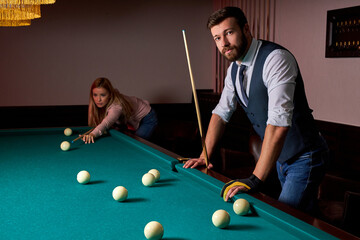 Fototapeta premium young active friends are playing billiards in bar after work, have rest and leaisure time, preparing aiming to shoot pool balls