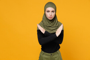 Displeased young arabian muslim woman in hijab black green clothes holding folded crossed hands in stop gesture isolated on yellow color background studio portrait. People religious lifestyle concept.