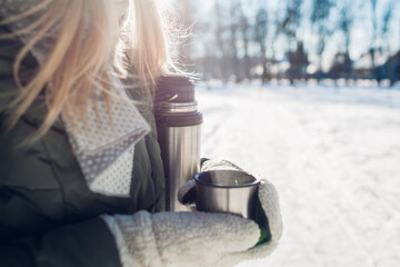 Woman drinking hot tea holding vacuum flask in winter park. Drinks to warm up in snowy frosty weather outdoors