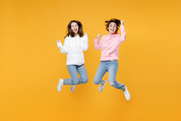 Fototapeta na wymiar Full length of excited two young women friends 20s in basic white pink hoodies jumping doing winner gesture celebrating clenching fists say yes isolated on yellow color background studio portrait.