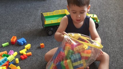 caucasian boy is picking up the constructor pieces and gathering them in a toy bag on the floor after playing