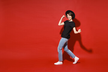 Fototapeta na wymiar Full length side view of joyful young brunette woman 20s in casual black t-shirt hat doing winner gesture celebrating clenching fists say yes isolated on bright red color background studio portrait.