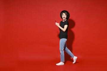 Fototapeta na wymiar Full length side view of shocked young brunette woman 20s in casual basic black t-shirt hat standing using mobile cell phone typing sms message isolated on bright red color background studio portrait.