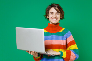 Smiling cheerful young brunette woman 20s years old in basic casual colorful sweater standing working on laptop pc computer looking camera isolated on bright green color background studio portrait.