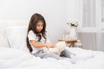 Obraz na płótnie Canvas sad little brunette girl sitting with teddy bear on bed at home