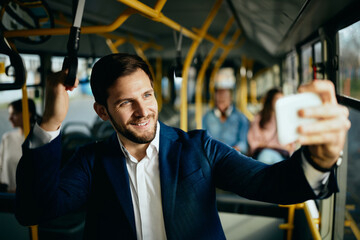 Happy businessman taking selfie while traveling to work by bus.