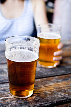 A caucasian woman, out of focus, holds a pint of beer at a picnic table.  In focus is another pint of beer that has been sipped on.