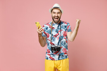 Happy traveler tourist man in summer clothes, hat using mobile phone typing sms message doing winner gesture isolated on pink background. Passenger traveling on weekends. Air flight journey concept.