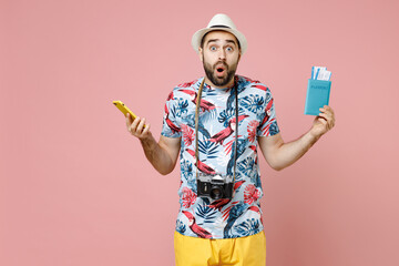 Shocked young traveler tourist man in summer clothes hat using mobile cell phone hold passport tickets isolated on pink background. Passenger traveling abroad on weekends. Air flight journey concept.