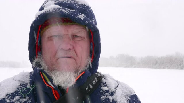 elderly man with gray beard stands on ice of snow-covered river under snowfall