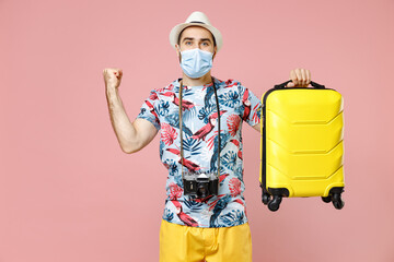 Joyful young traveler tourist man in sterile mask to safe from coronavirus hold suitcase doing winner gesture isolated on pink background. Passenger traveling on weekend. Air flight journey concept.