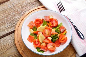 Salad with salmon and cherry tomatoes and green salad in a plate on a wooden table on a wooden stand stand next to a fork.