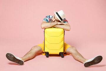 Full length of tired young traveler tourist man in summer clothes hat sit on floor sleeping on suitcase isolated on pink background. Passenger traveling abroad on weekend. Air flight journey concept.