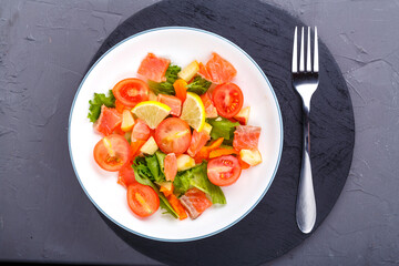 Salad with cold salmon and cherry tomatoes in a plate on a concrete background with a black stand next to a fork.