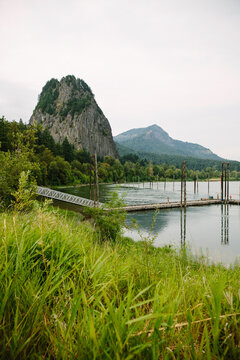 A dock stretches out over the water with a view of mountains and foliage in the background and tall grass in the foreground.