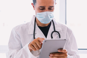 Black haired doctor in white coat and stethoscope looking at his tablet, in a window. Medicine concept