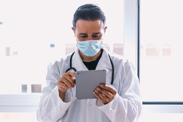Black haired doctor in white coat and stethoscope looking at his tablet, in a window. Medicine concept