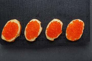 Bruschetta with butter and red caviar and a spoon with caviar on a black board in a row. copy space.