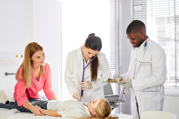 Fototapeta premium team of doctors working together while treating kid girl patient lying on hospital bed, african man and caucasian woman consulting girl and holding medical check-up