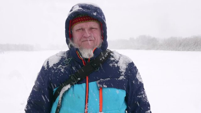 elderly man with gray beard stands on ice of snow-covered river under snowfall