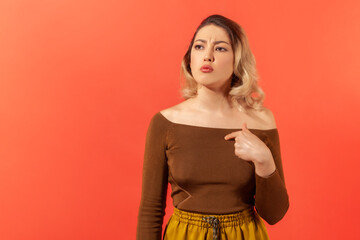 Portrait of serious young woman in casual clothes pointing at herself by finger and can't believe it's really true. Sudden sucsess and good fortune. Indoor studio shot isolated on red background
