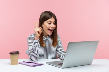 Smiling positive woman office worker in striped shirt pointing finger up talking video call on laptop, great idea for startup, business solution. Indoor studio shot isolated on pink background