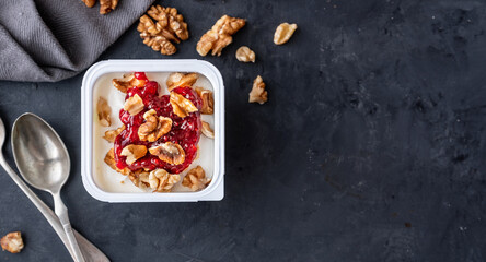 White yogurt in plastic square cup with marmalade and spoon in with walnuts on gray background....