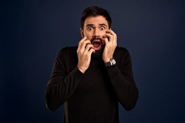 Studio shot of shocked scared man being in panic, keeps hands near opened mouth, sees something terrible, feels excited, wears black sweater sweatshirt, isolated on blue background, hears awful news