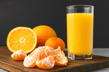 Glass of fresh orange juice, whole and sliced fruits on wooden board on dark background with copy-space