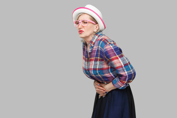 Stomach pain. Profile side view portrait of modern stylish mature woman in casual style with hat and eyeglasses standing and holding her painful belly. indoor studio shot isolated on gray background