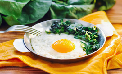 Fried egg with spinach on iron pan plate. Fried eggs healthy breakfast on wooden table background served by orange napkin, fork and spinach leaves, ingredients. Close up