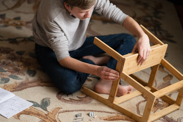 Teen boy assembles furniture according to the instructions - a wooden chair. Housework and order in it. Lesson for a teenage boy on vacation. Assistant.