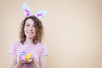 Obraz na płótnie Canvas Pretty smiling girl in bunny ears, striped pink t-shirt is holding eggs. Young curly woman is preparing for celebration. Happy easter, spring concept. Carnival, seasonal party decor for holiday.