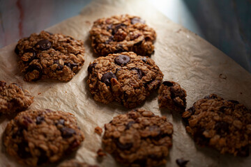 Angled view of fresh made cookies on baking paper. These are made with whole grain rolled oats with dark chocolate chips. There are pieces and crumbles on the paper. Lighting is directional and moody.