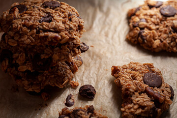 Stacked up fresh made cookies on baking paper. These are made with whole grain rolled oats with dark chocolate chips. There are pieces and crumbles on the paper. Lighting is directional and moody.