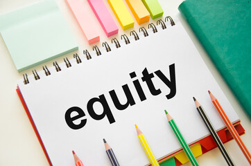 Word EQUITY written on a white notebook that lies on a white table near stickers and colored pencils